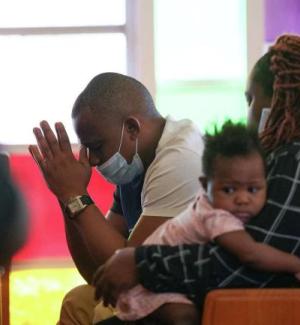 Portrait of an African American man, woman and baby sitting on a church pew at Boynton Chapel United Methodist Church. The man is leaning forward, with hands in prayer position. The woman sits beside him, looking towards the man, while the baby in her arms looks into the camera.