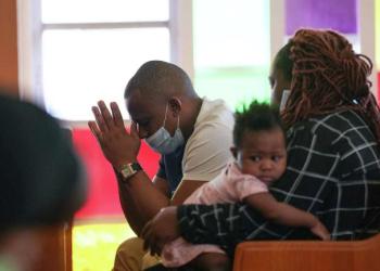 Portrait of an African American man, woman and baby sitting on a church pew at Boynton Chapel United Methodist Church. The man is leaning forward, with hands in prayer position. The woman sits beside him, looking towards the man, while the baby in her arms looks into the camera.