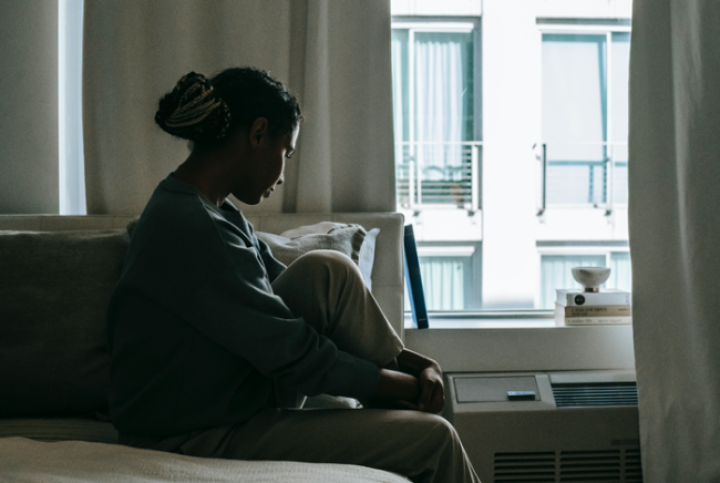 Photo by Alex Green from Pexels. Photo shows woman in an apartment sitting on bed looking down towards a window. The lighting is bright outside but dark indoors.