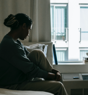 Photo by Alex Green from Pexels. Photo shows woman in an apartment sitting on bed looking down towards a window. The lighting is bright outside but dark indoors.
